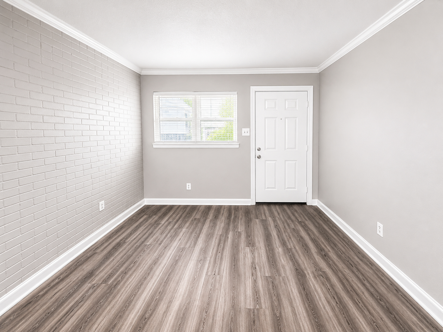 An empty room with light gray walls, a white door on the right, and a window. The floor is covered with brown laminate flooring.