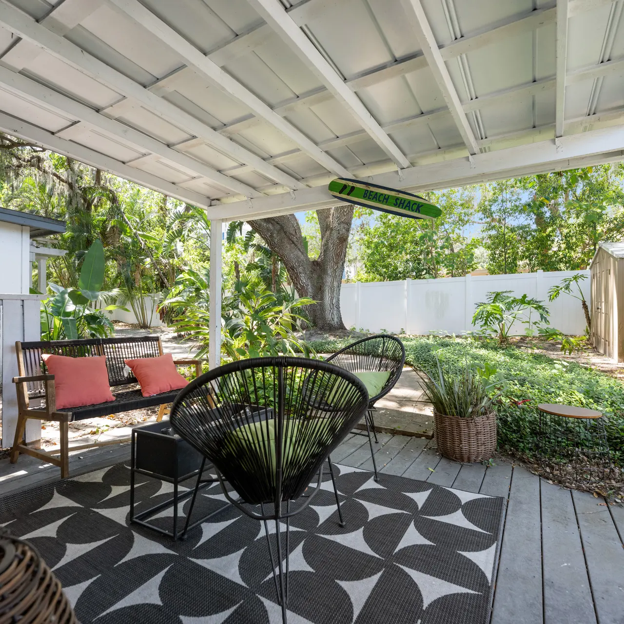 Cozy outdoor patio area with black and white patterned rug, two seating options including a black chair and wooden bench, surrounded by greenery.