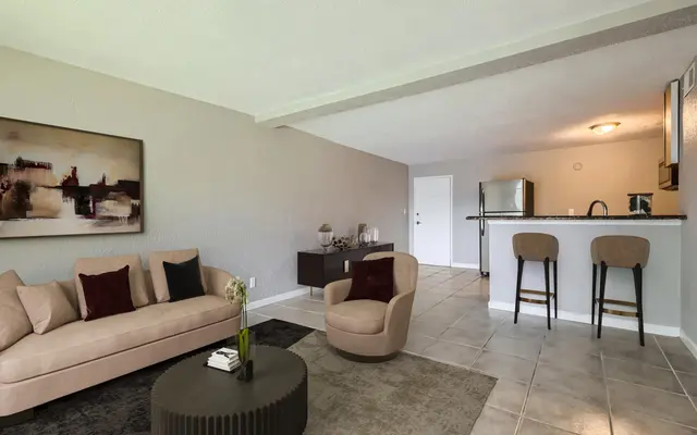 A modern living room featuring a sofa with cushions, a round coffee table, and two armchairs. The space has neutral-colored walls and tile flooring, with a kitchenette in the background.
