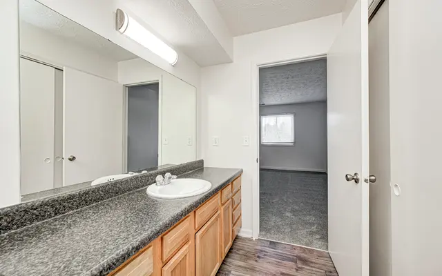 A spacious bathroom with a double sink vanity, large mirror, and wooden cabinets. A doorway leads to another room with a window.