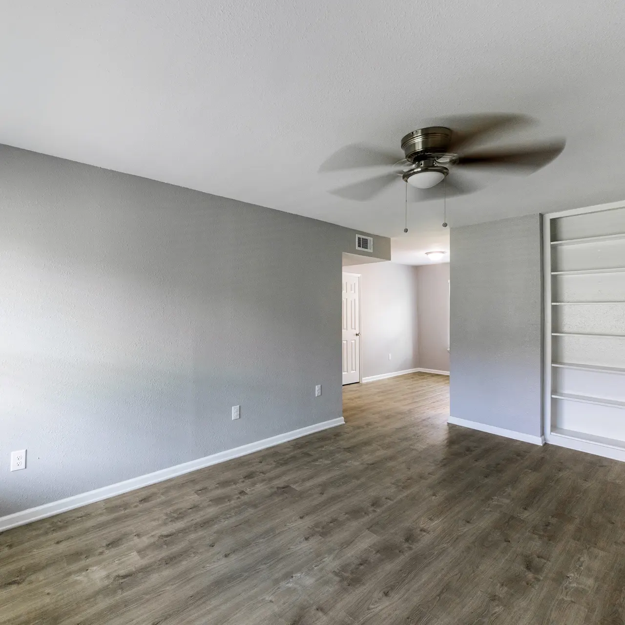 An empty room with gray walls and a wooden floor, showcasing a ceiling fan and a built-in shelf.