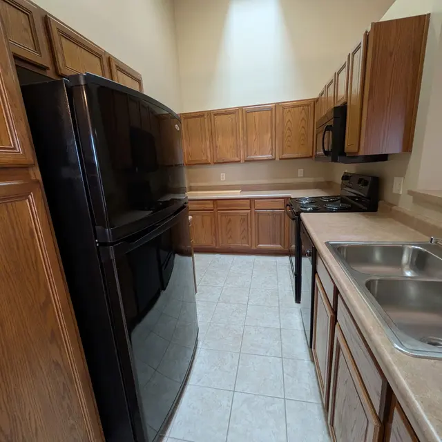 A modern kitchen featuring wooden cabinets, a black refrigerator, a stove, and a double sink. The kitchen has tile flooring and a skylight for natural light.
