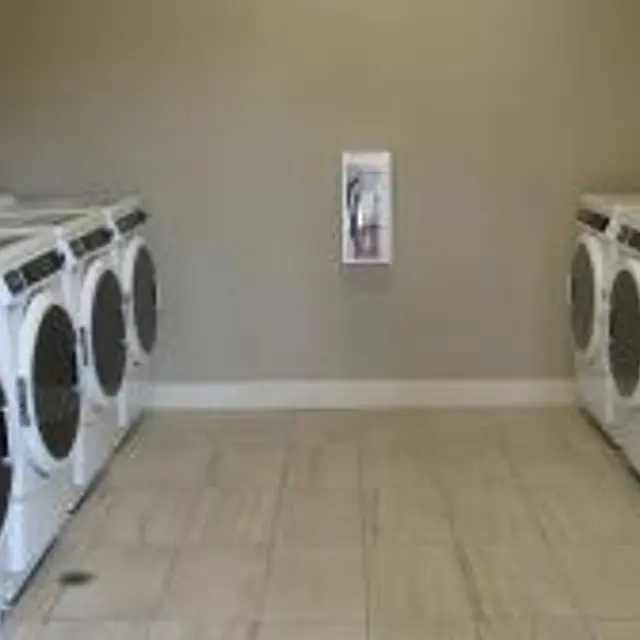 A clean and spacious laundry room with several washers and dryers lined up against the walls, tiled flooring, and plain walls.