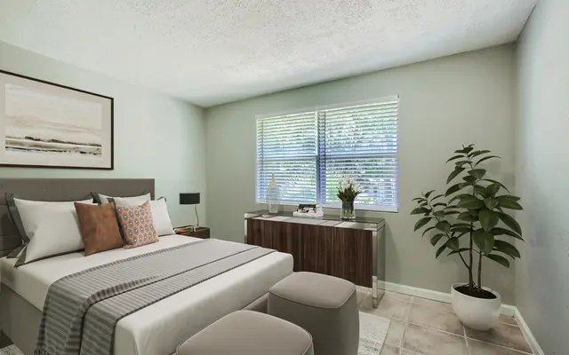 A modern bedroom featuring a double bed with decorative pillows, a cozy gray throw blanket, and two upholstered stools. A wooden dresser can be seen against the wall beneath a window with blinds, alongside a potted plant. The walls are painted light green and the flooring is tiled.