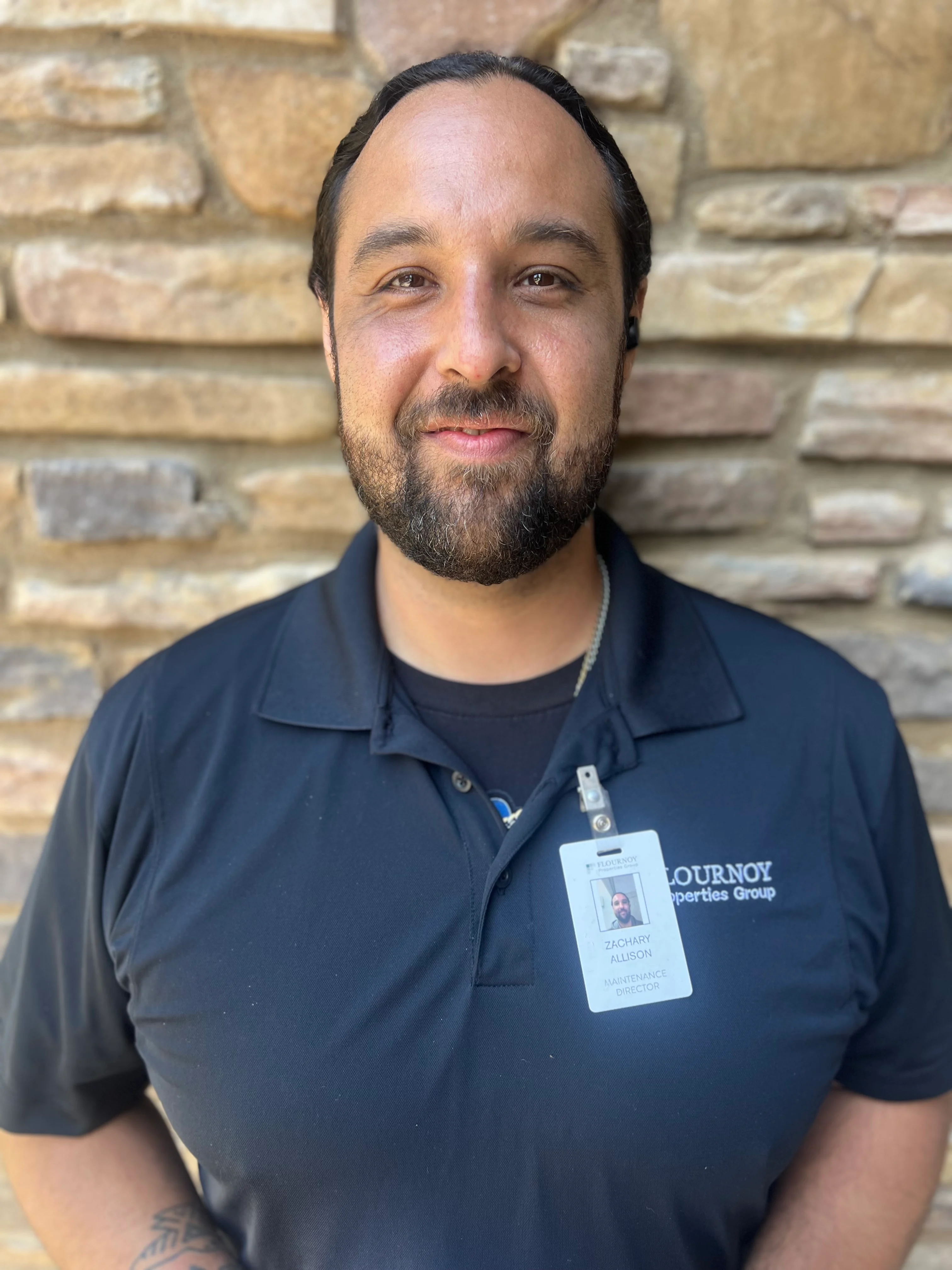 A man with a beard wearing a black polo shirt with a name badge, standing in front of a stone wall.