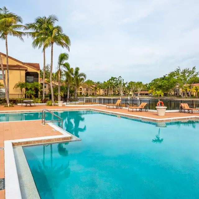 A tranquil swimming pool area with palm trees and lounge chairs, surrounded by residential buildings and water.
