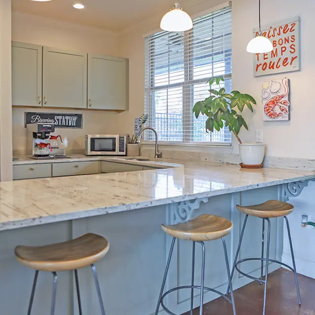 A modern kitchen interior with a light marble countertop, bar stools, and large windows. There’s a potted plant near the window and kitchen appliances in view.