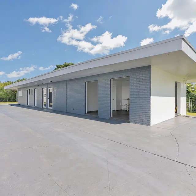 Side view of a modern one-story leasing office with a gray brick façade and large windows, surrounded by greenery and blue skies