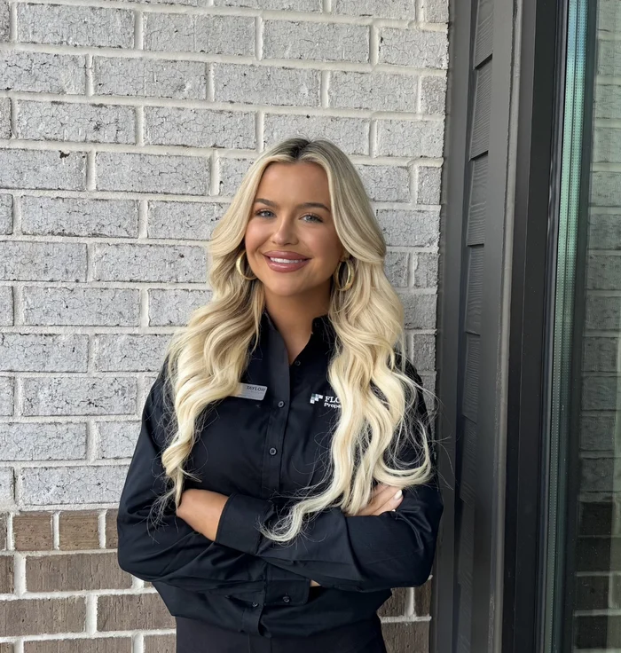 A smiling young woman with long blonde hair, wearing a black button-up shirt, stands with her arms crossed against a brick wall.