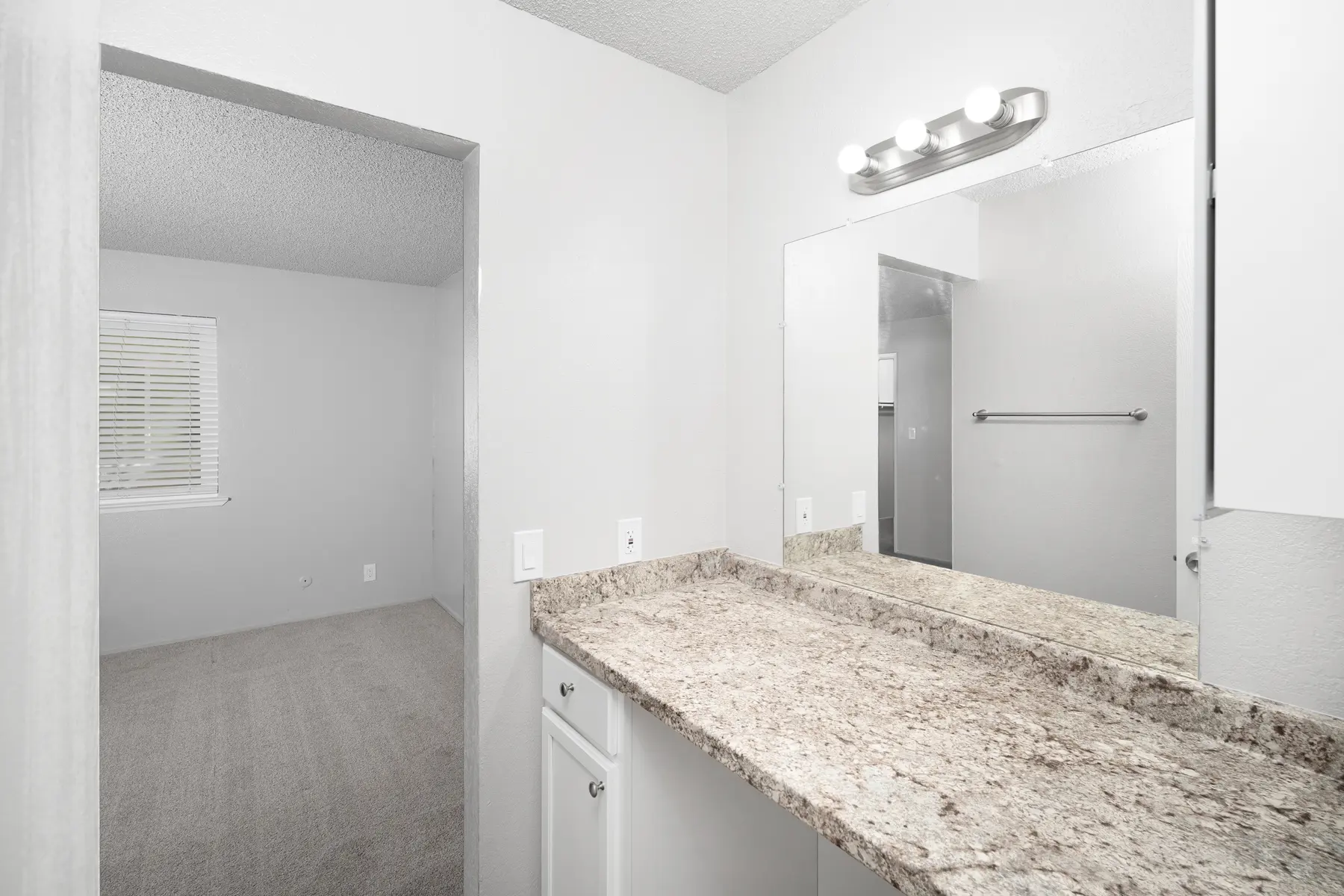 A modern bathroom view featuring a granite countertop, a mirror, and a glimpse of an adjacent room with a window.