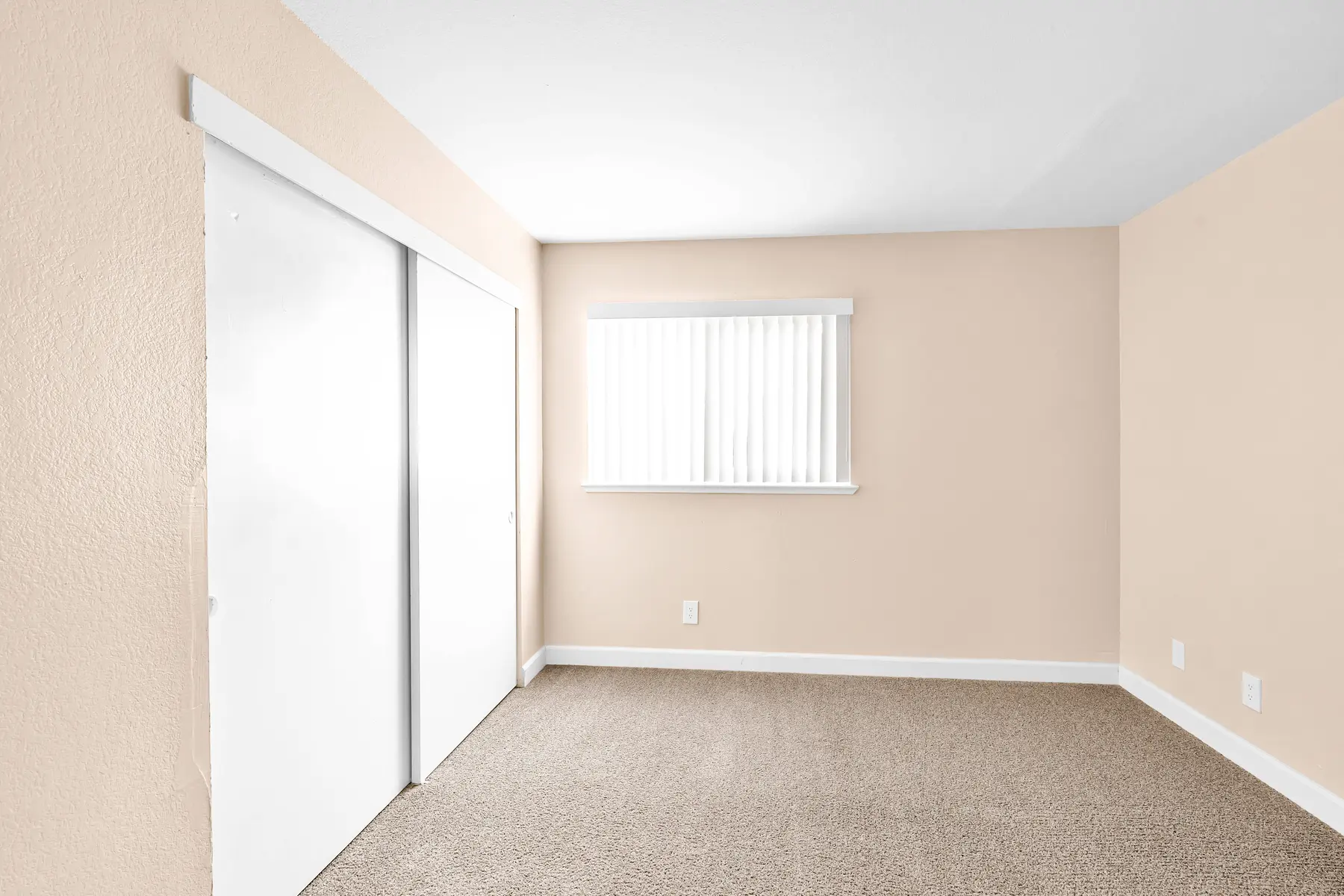 A vacant room with beige walls, carpet flooring, a window with white blinds, and sliding closet doors.