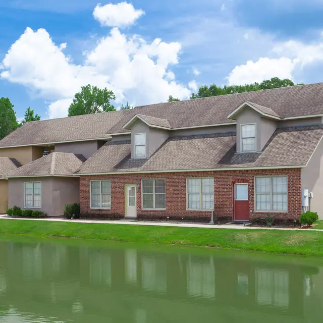 An image of a residential building beside a pond, featuring brick and stucco architecture, surrounded by greenery and blue skies.