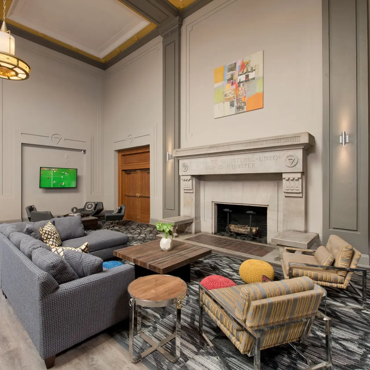 A spacious modern living room featuring a gray sectional sofa, colorful accent chairs, a coffee table, and a stone fireplace. A wall-mounted television displays a program. Natural light streams in through large windows, highlighting the high ceilings and decorative moldings.