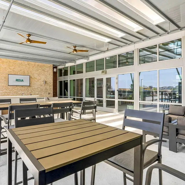 An outdoor seating area with modern furniture, including wooden tables and metal chairs, under a spacious covered structure with large windows.