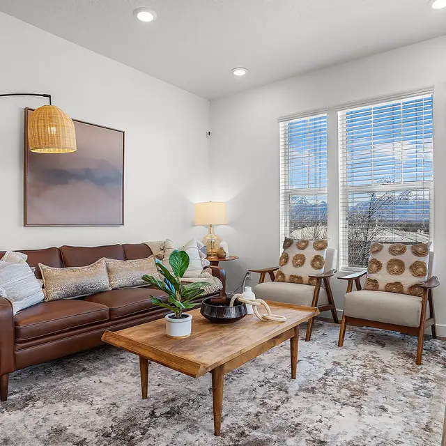 A cozy living room featuring a brown leather sofa, a wooden coffee table, and contemporary decor. Large windows allow natural light to fill the space, complemented by modern lamps and a vibrant rug.