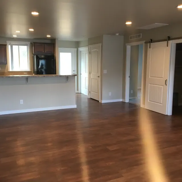 Spacious Living Area A spacious living area featuring hardwood floors, bright lighting, a kitchenette with dark cabinetry, and two doors leading to other rooms.