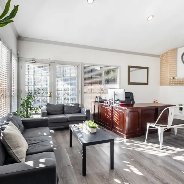A bright and modern office/living room space featuring a comfortable seating area with dark couches and a black coffee table. A desk is positioned in the background against a wall with a clock, and there are plants near the windows, creating a cozy atmosphere.