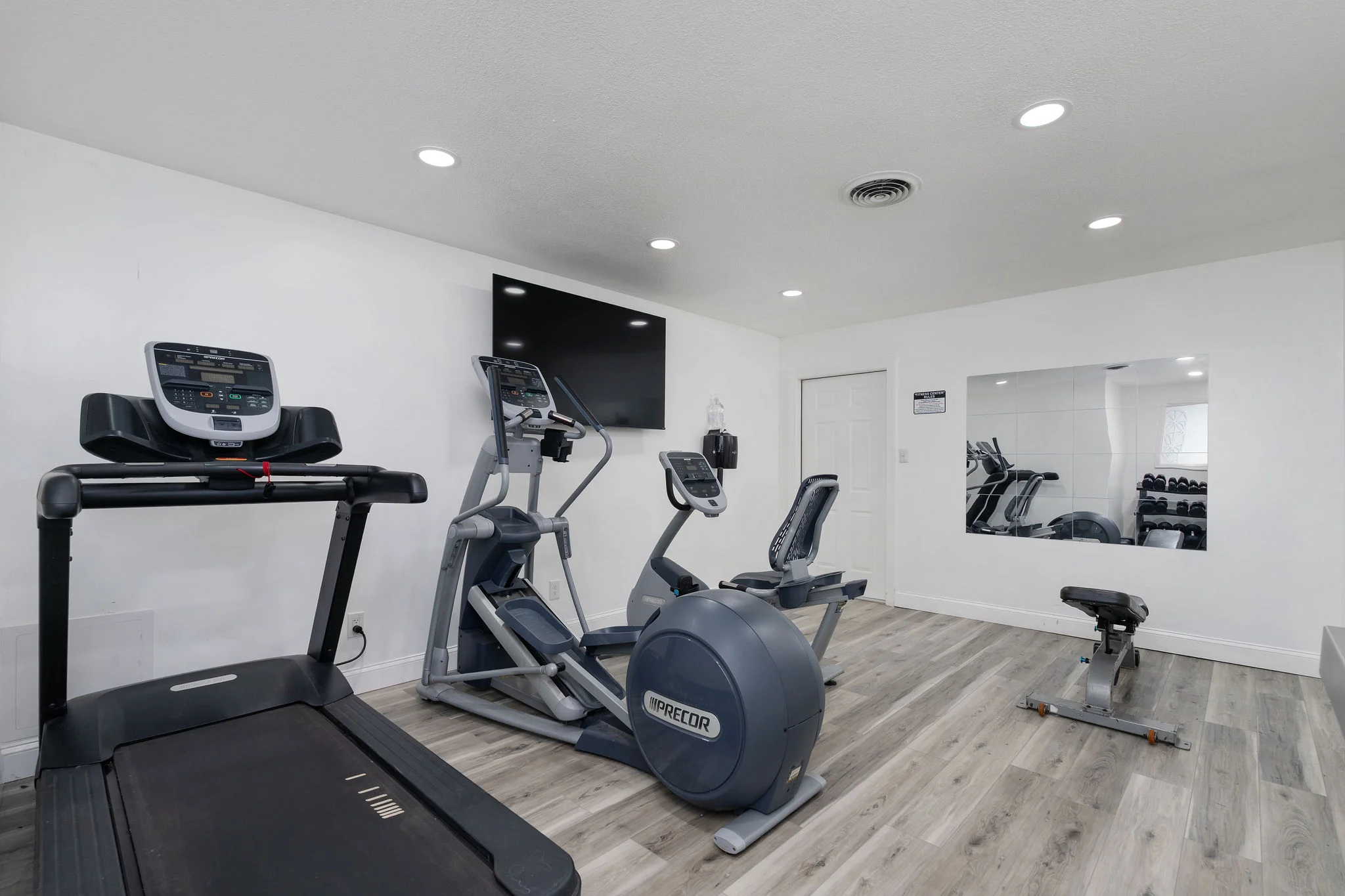 Home Gym Setup A modern home gym featuring a treadmill, elliptical machine, exercise bike, and weight bench. Brightly lit with white walls and flooring.