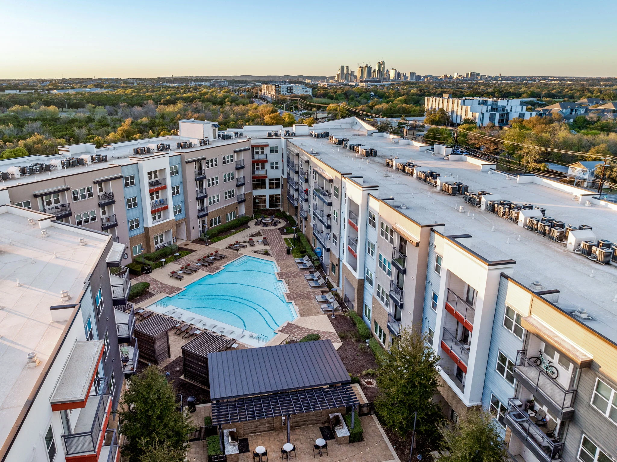 Aerial view of a modern apartment complex with a pool in the center, surrounded by green trees and the city skyline in the background.