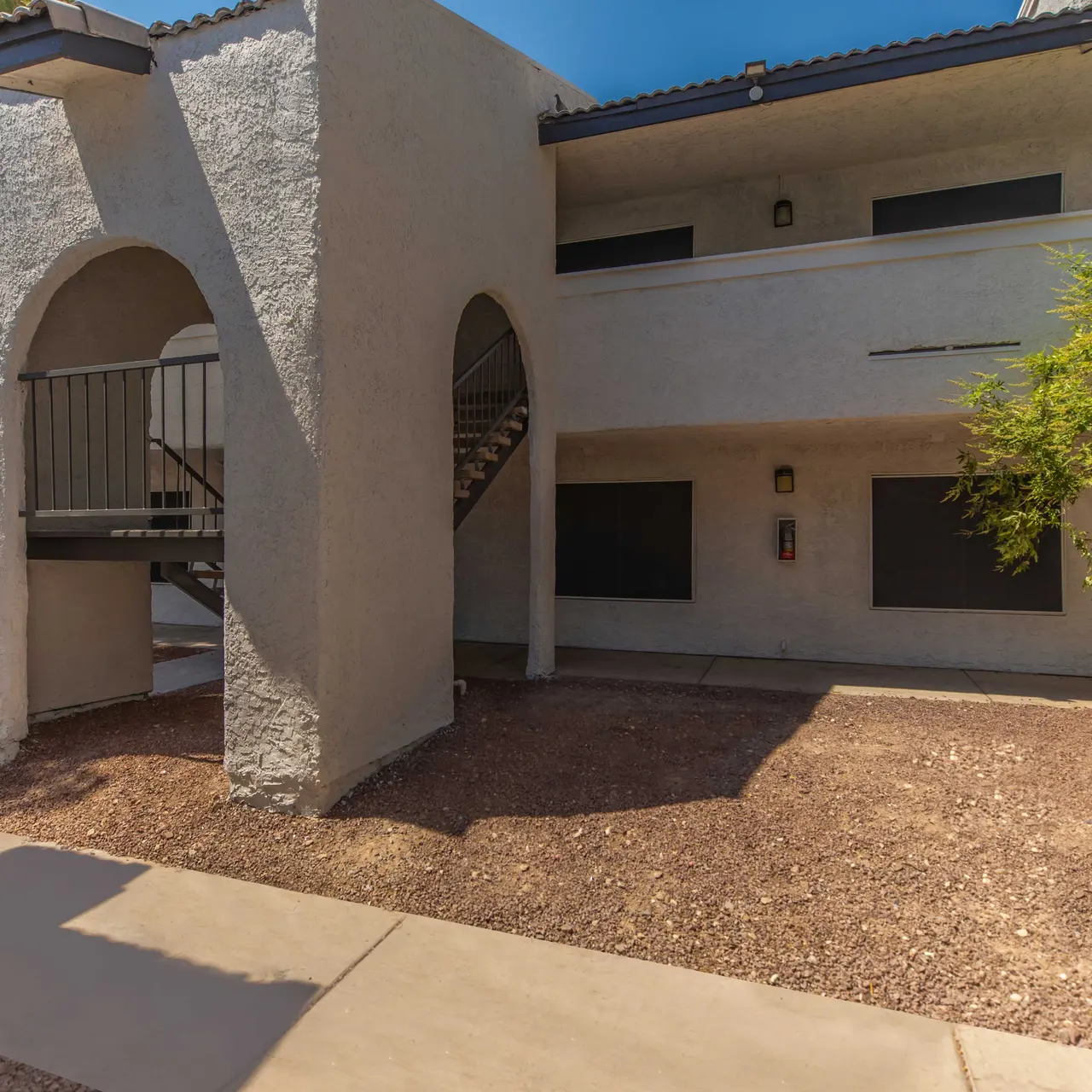Exterior view of an apartment complex with an archway and staircase, surrounded by gravel and landscaping.
