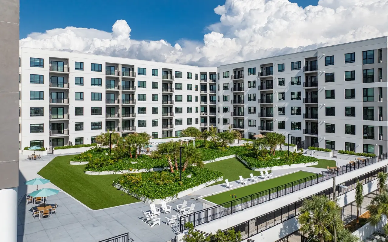 A modern apartment complex featuring multiple floors with large windows, surrounded by landscaped green areas and palm trees.