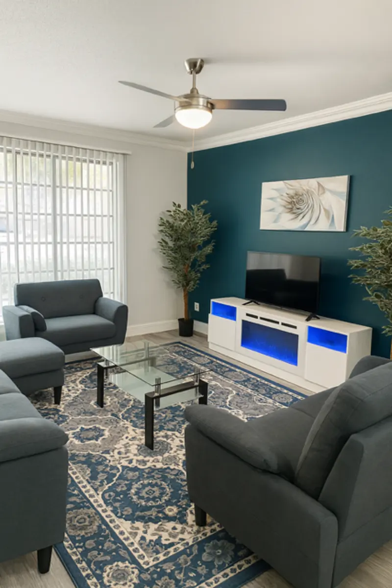 A modern living room featuring two gray armchairs and a matching couch, a glass coffee table, a TV stand with blue highlights, and two green plants. The wall is painted dark blue and there are large window blinds letting in natural light.