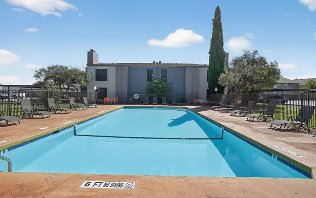 A swimming pool surrounded by lounge chairs and greenery at an apartment complex on a sunny day.