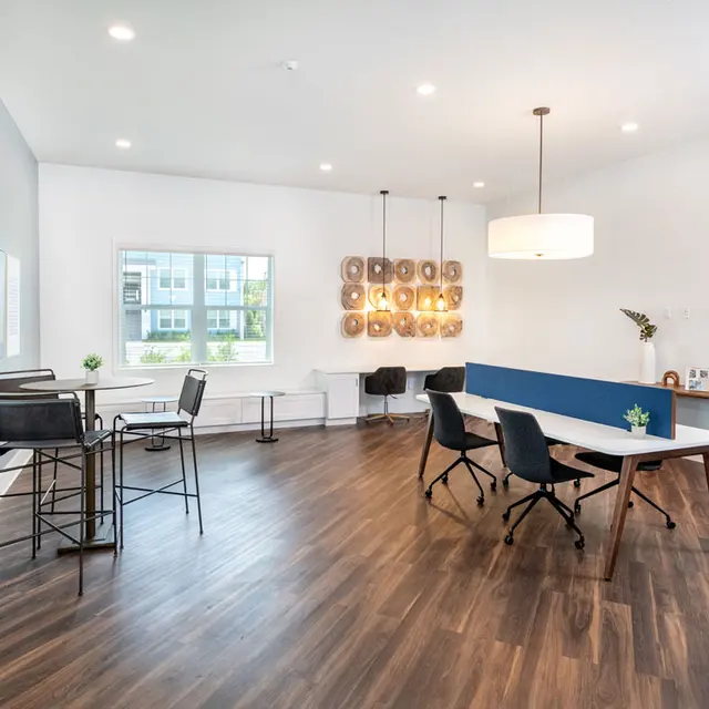 A modern office space featuring a large table, black chairs, and a stylish light fixture. The room has wood flooring and bright natural light coming from windows.