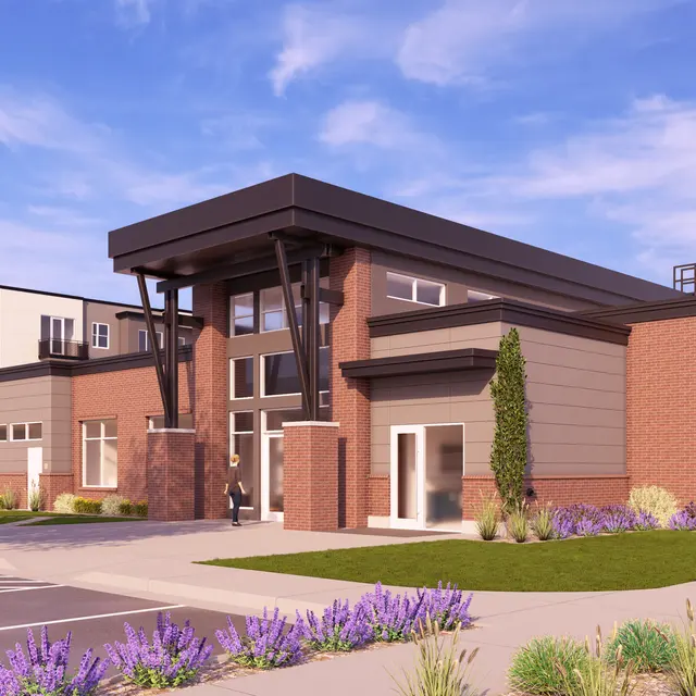 A modern building with a brick facade and large glass windows, surrounded by greenery and flowering plants. The entrance features a large overhang supported by wooden beams. In the background, additional buildings are visible, along with parked cars.