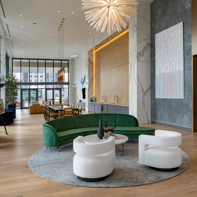A spacious and modern lobby with a green curved sofa and white chairs, featuring large windows with natural light, wood flooring, and contemporary decor elements including plants and artwork.
