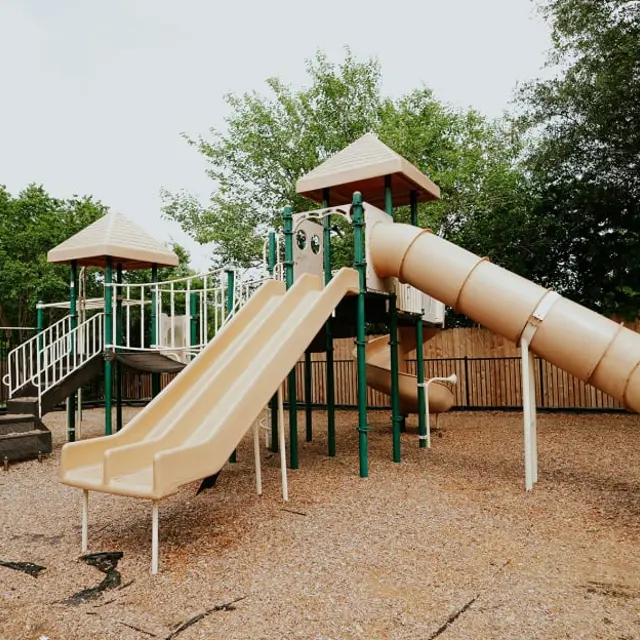 A playground featuring two slides and a spiral slide, set in a gravel area surrounded by green trees and a safety fence.