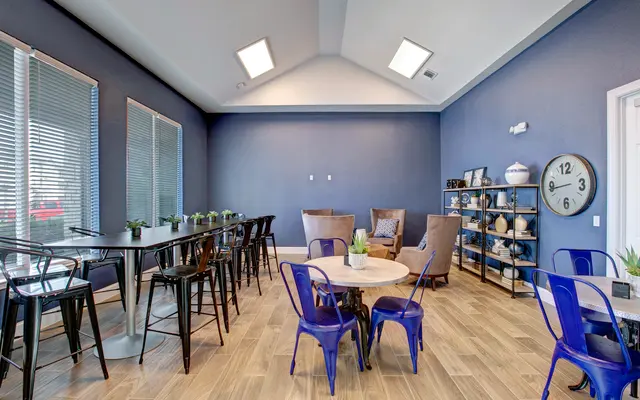 A modern lounge area with a blue wall, featuring a mix of seating options including high tables with black chairs and a cozy seating arrangement with brown armchairs. Decorative plants and a wall clock are visible.