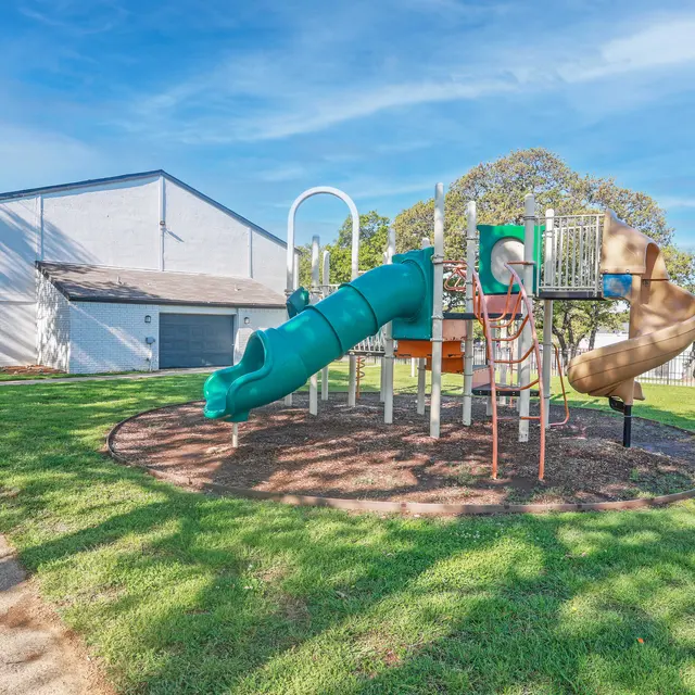 A playground featuring a green slide and a tan slide, set on a grassy area next to a building.