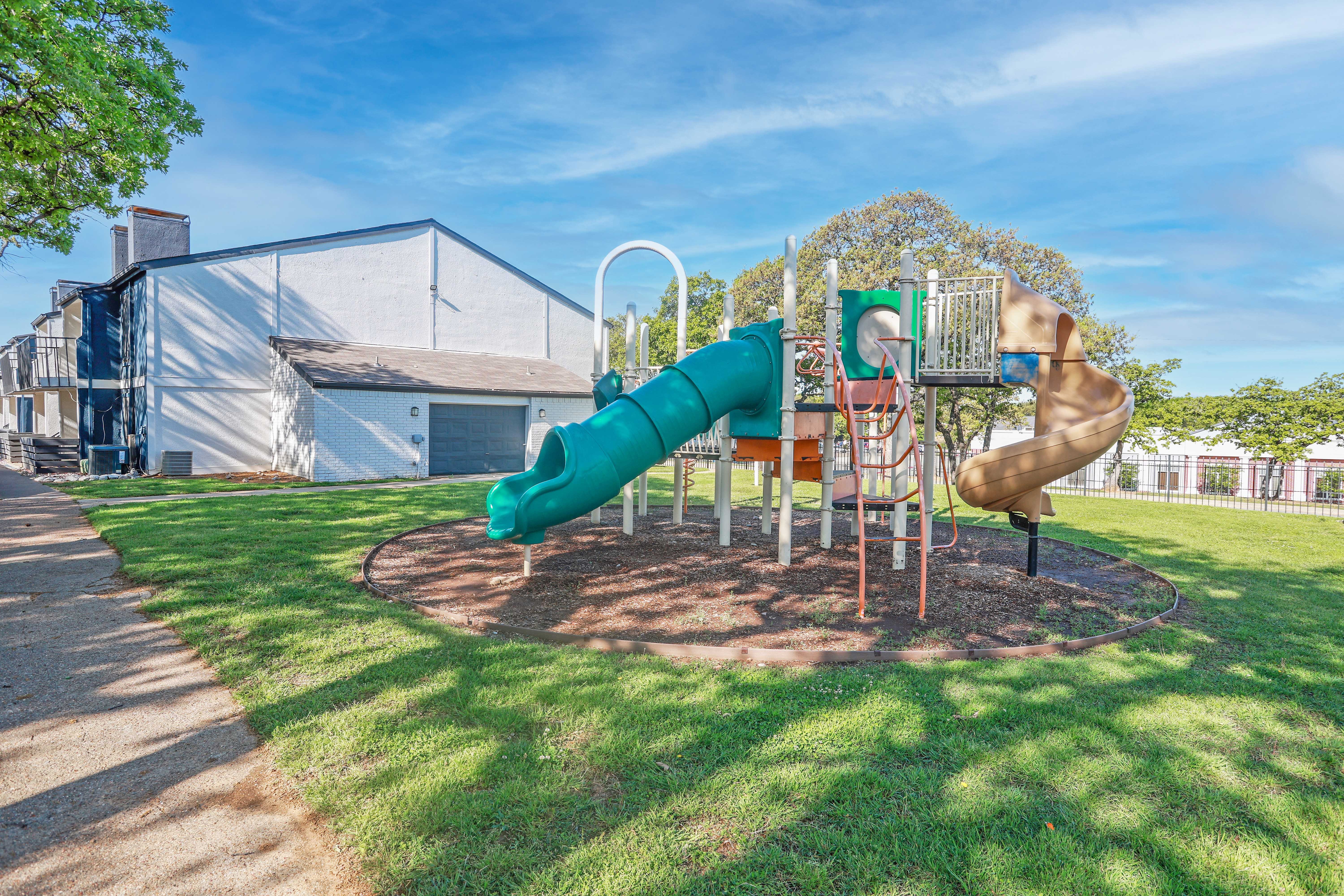 A playground featuring a green slide and a tan slide, set on a grassy area next to a building.