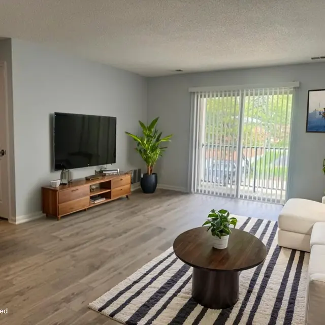 A modern living room with a white sofa, a round wooden coffee table, and a large TV on a wooden cabinet. There is a plant next to the sofa and large sliding glass doors leading outside. The floor is light wood with a striped rug.