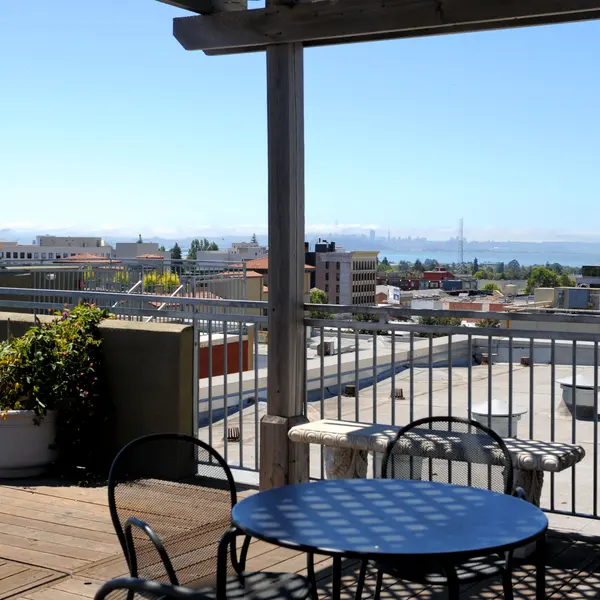 A panoramic view from a rooftop terrace featuring a round table and chairs, overlooking a cityscape with distant mountains.