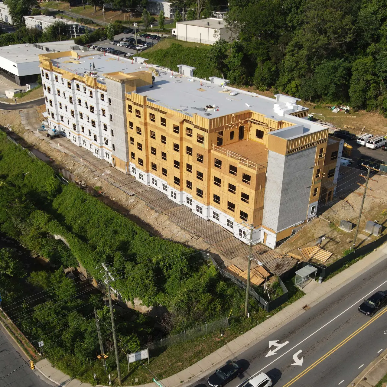 Aerial view of a new multi-story building under construction, featuring wooden frames and a partially finished exterior. Surrounding greenery and roads are visible.