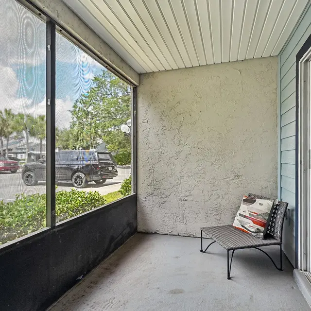 Charming Screened Patio A screened patio area with a small chair and a decorative pillow, overlooking a green garden and a view of parked cars.