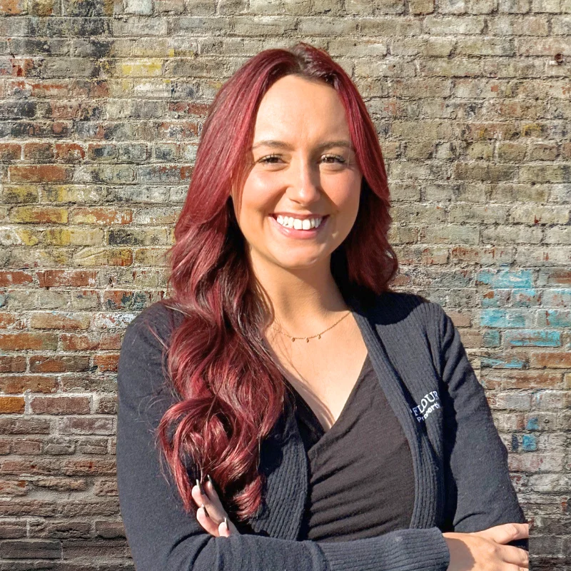 A woman with long red hair smiles confidently, standing with her arms crossed against a brick wall.