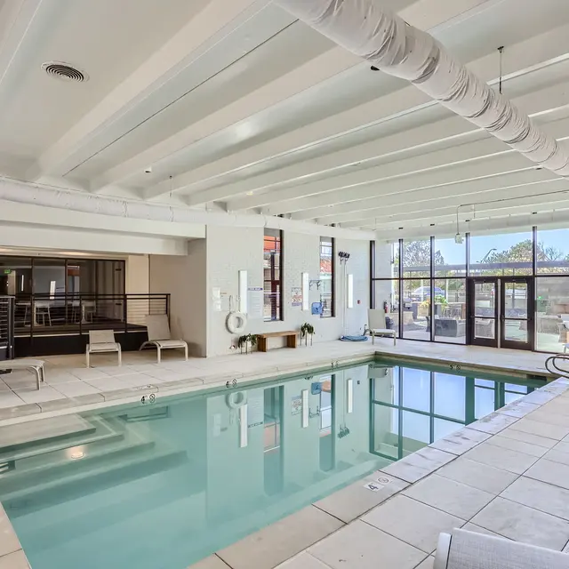 A modern indoor pool area with large windows and natural light. The pool features a tiled perimeter and is surrounded by lounge chairs. A spacious deck with a contemporary design complements the swimming area, and there are plants and amenities visible.