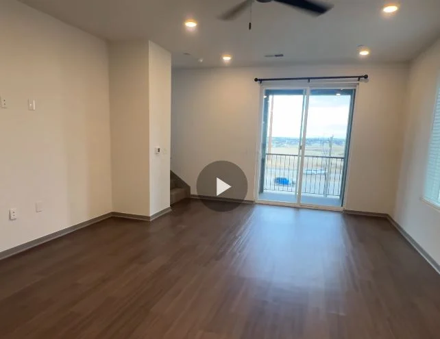 A spacious modern living room with wooden floors, large windows, and a balcony. The walls are light-colored, and there is a ceiling fan with lights.