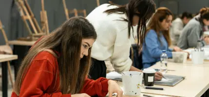 Students engaged in an art class, focused on their work at wooden tables with art supplies.