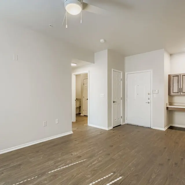 A spacious apartment interior with light-colored walls and wooden flooring. The room includes a ceiling fan, a small kitchenette area, and entrances to other rooms.