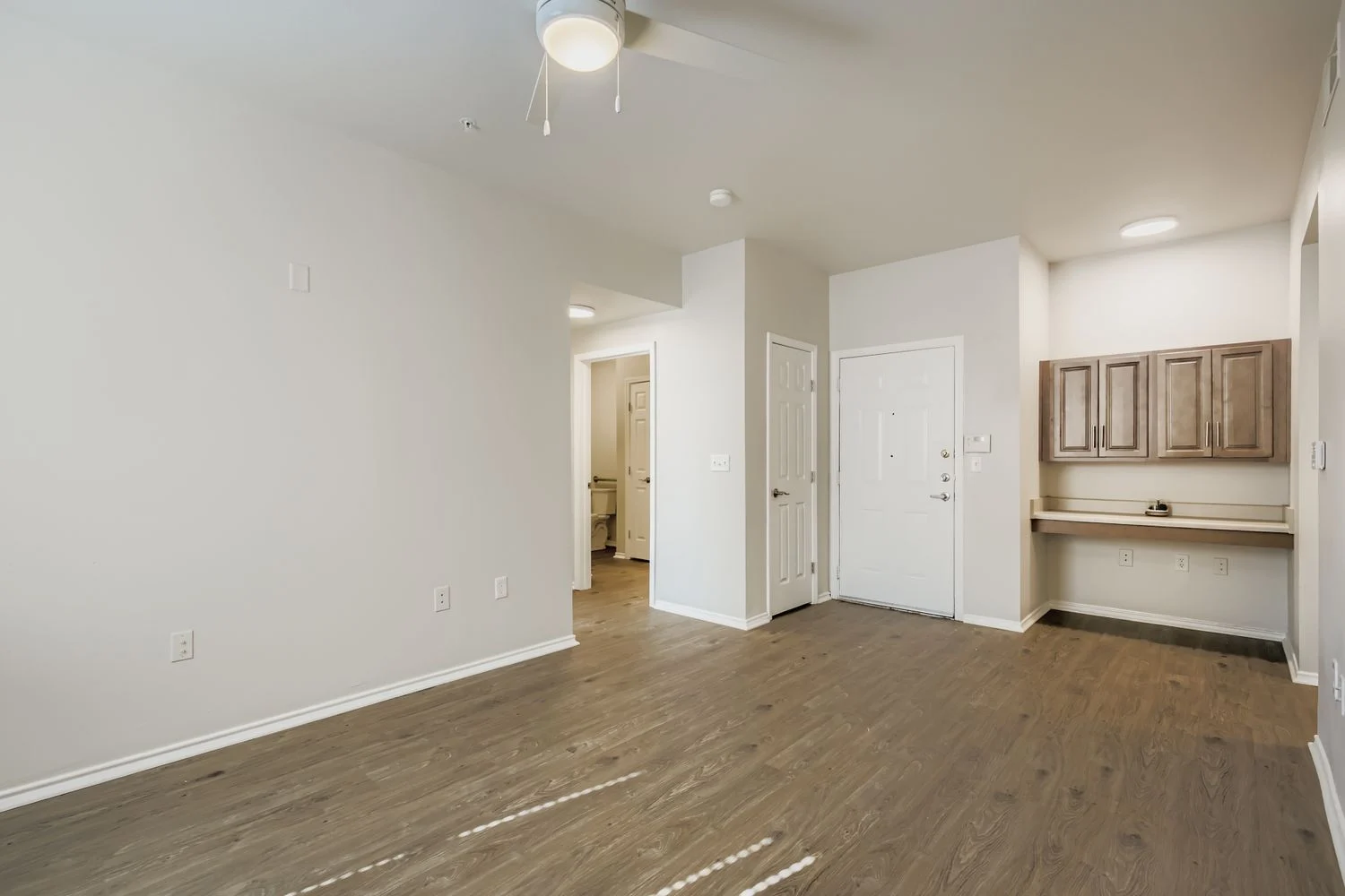 Modern Apartment Interior A spacious apartment interior with light-colored walls and wooden flooring. The room includes a ceiling fan, a small kitchenette area, and entrances to other rooms.