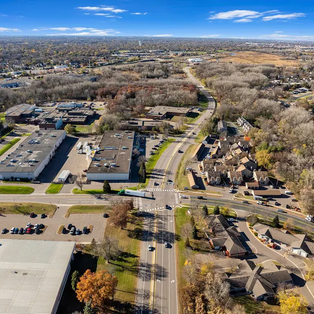 Aerial view of a suburban area showcasing a mix of residential houses and commercial buildings, surrounded by trees and open fields under a clear blue sky.