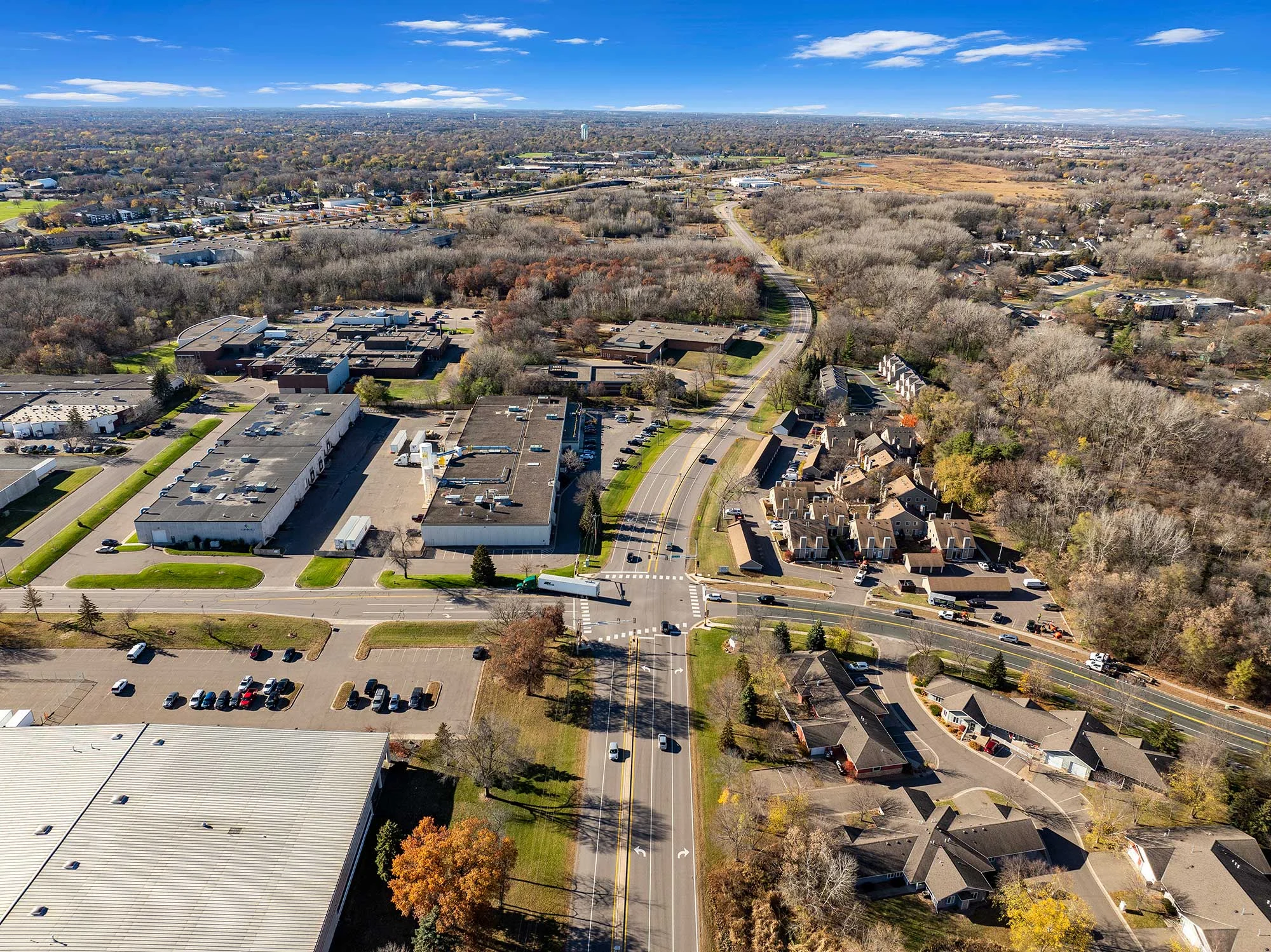 Aerial View of Suburban Area Aerial view of a suburban area showcasing a mix of residential houses and commercial buildings, surrounded by trees and open fields under a clear blue sky.