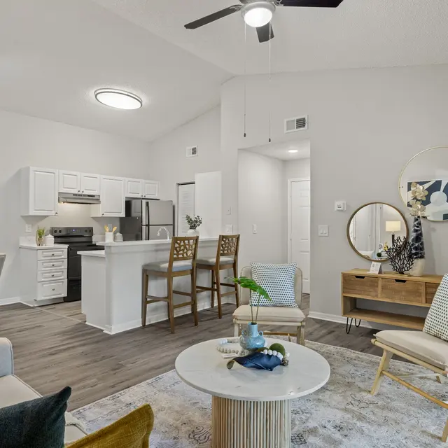 A modern living space featuring a dining area and kitchen with white cabinets, a stylish round coffee table in front, and comfortable seating with decorative pillows. The room has a high ceiling, a ceiling fan, and a large wall painting.