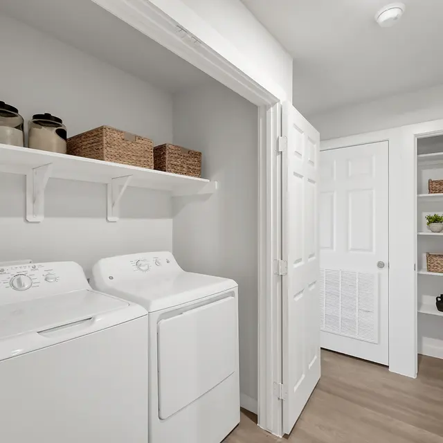 A modern laundry room featuring a washing machine and dryer side by side, white shelves above with decorative containers and baskets, and a door leading to a storage area with shelves.