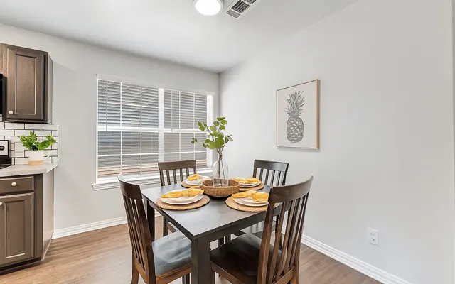 A cozy dining setup featuring a black table with four wooden chairs, set with yellow plates. A vase with green plants sits at the center. The kitchen area is visible in the background, featuring dark cabinets and a window with white blinds.
