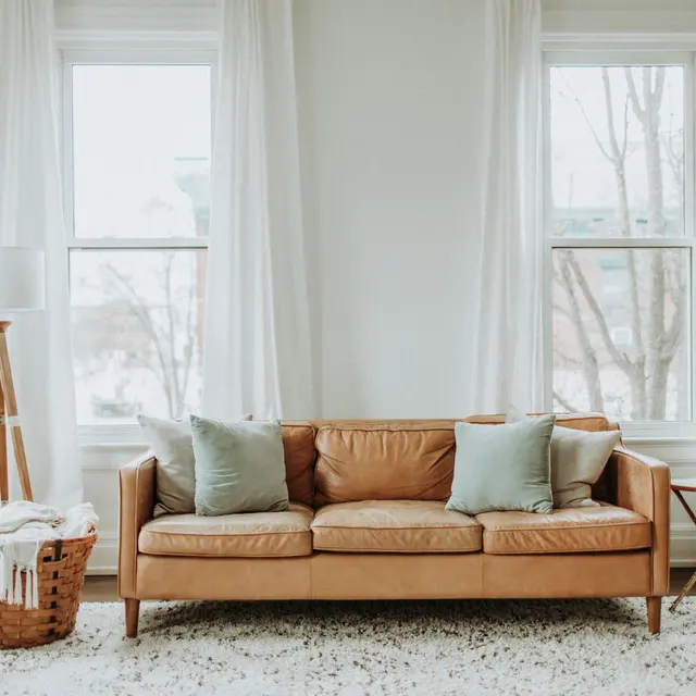 A cozy living room featuring a tan leather sofa with pillows, a floor lamp, a decorative basket, and windows draped with white curtains.